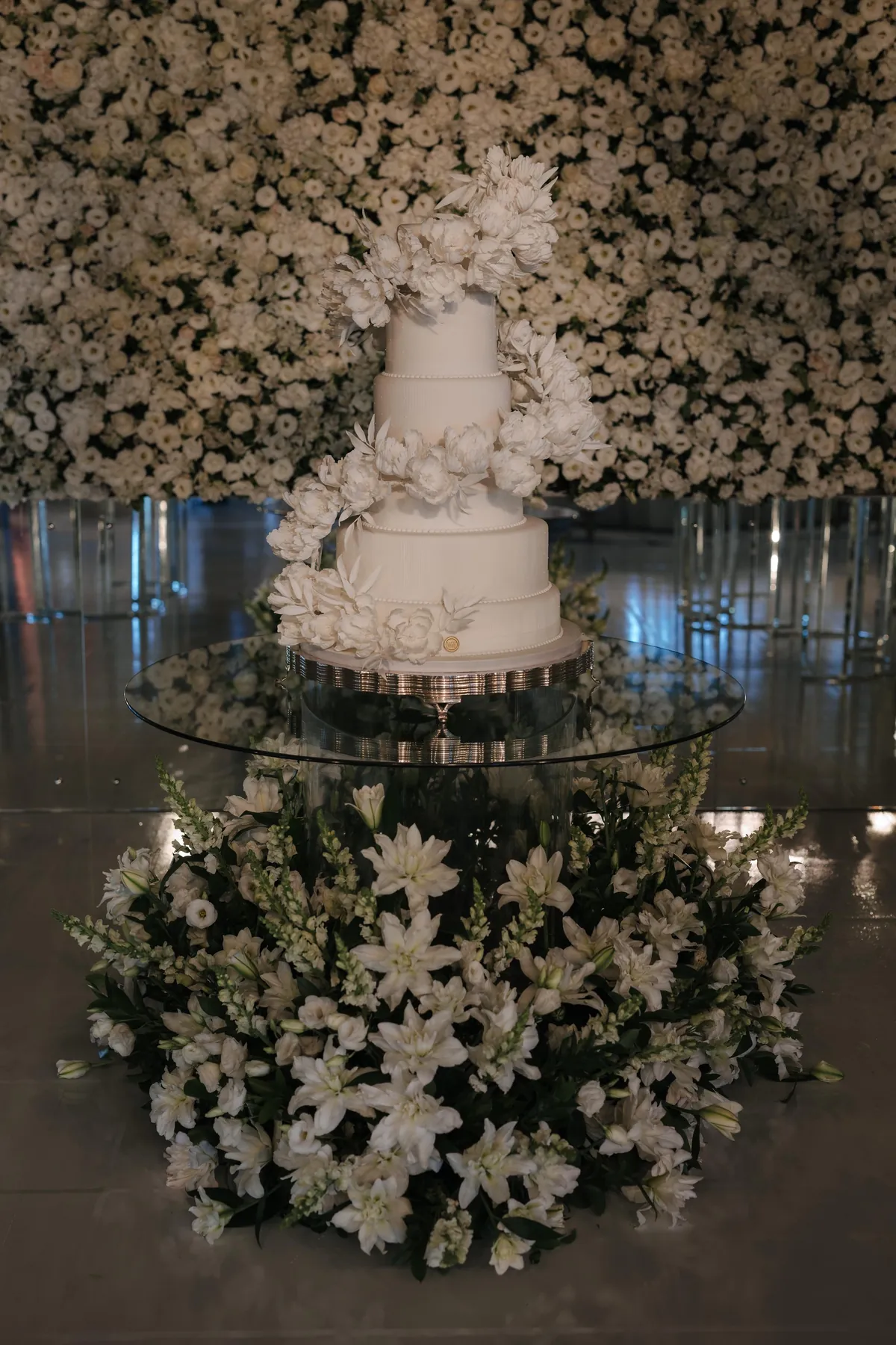 Bolo branco sobre mesa de vidro diante de um paredão de flores brancas