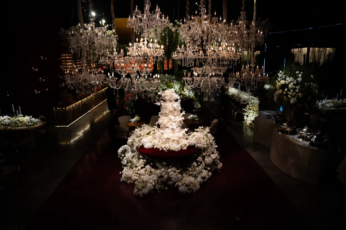 Bolo branco de flores sob dezenas de lustres de cristal, em visão ampla do salão
